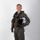 MCAS Marines pose for content 2026 A young male military aviator stands in a full-body portrait pose against a plain white/light grey studio background. He is wearing a complete flight suit ensemble including a green/olive flight suit, survival vest, harness, and neck collar, and is holding a white flight helmet under his arm. The image appears to be an official or professional military portrait photograph.