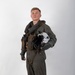 A young male military aviator stands in a full-body portrait pose against a plain white/light grey studio background. He is wearing a complete flight suit ensemble including a green/olive flight suit, survival vest, harness, and neck collar, and is holding a white flight helmet under his arm. The image appears to be an official or professional military portrait photograph.