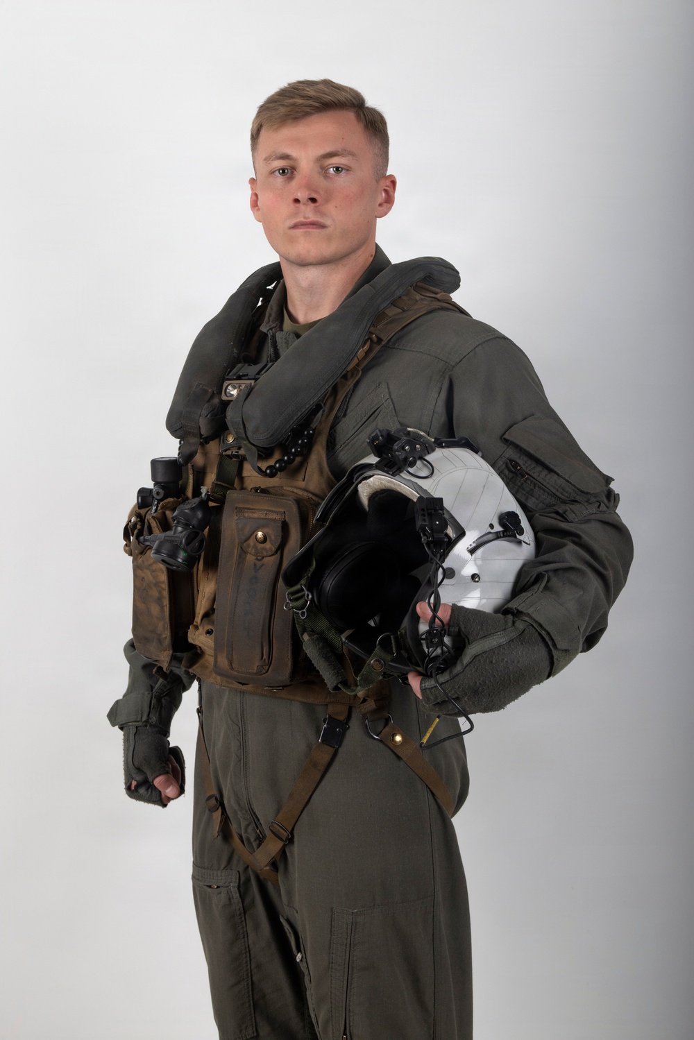A young male military aviator is posed against a plain white/grey studio background in a formal portrait-style photograph. He is wearing a full flight suit ensemble with survival/tactical gear and is holding a flight helmet under his arm. The image appears to be an official or professional military portrait.