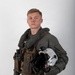 A young male military aviator is posed against a plain white/grey studio background in a formal portrait-style photograph. He is wearing a full flight suit ensemble with survival/tactical gear and is holding a flight helmet under his arm. The image appears to be an official or professional military portrait.