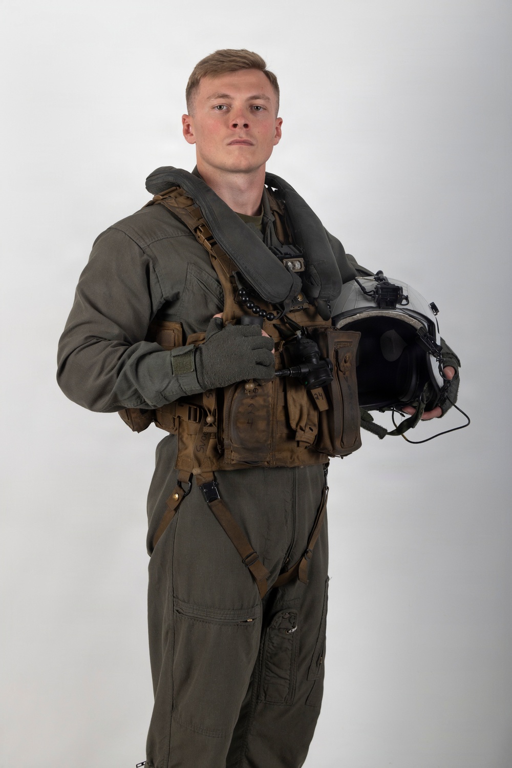 A young male military aviator poses in a professional portrait-style photograph against a neutral gray-white background. He is wearing a full flight suit ensemble including a green/olive flight suit, survival vest, harness, and anti-exposure collar, and is holding a flight helmet in his right hand. The image appears to be an official or semi-official military portrait.