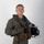 MCAS Marines pose for content 2026 A young male military aviator poses in a professional portrait-style photograph against a neutral gray-white background. He is wearing a full flight suit ensemble including a green/olive flight suit, survival vest, harness, and anti-exposure collar, and is holding a flight helmet in his right hand. The image appears to be an official or semi-official military portrait.