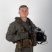A young male military aviator poses in a professional portrait-style photograph against a neutral gray-white background. He is wearing a full flight suit ensemble including a green/olive flight suit, survival vest, harness, and anti-exposure collar, and is holding a flight helmet in his right hand. The image appears to be an official or semi-official military portrait.