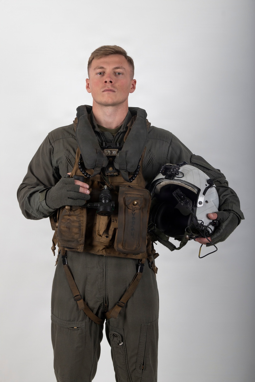 A young male military aviator poses for a formal portrait against a neutral gray-white studio background. He is wearing a full flight suit ensemble including a green flight suit, survival vest, harness, and partial-finger tactical gloves. He holds a flight helmet in his left hand and an oxygen mask assembly in his right hand.