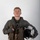 MCAS Marines pose for content 2026 A young male military aviator poses for a formal portrait against a neutral gray-white studio background. He is wearing a full flight suit ensemble including a green flight suit, survival vest, harness, and partial-finger tactical gloves. He holds a flight helmet in his left hand and an oxygen mask assembly in his right hand.
