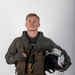 A young male military aviator poses for a formal portrait against a neutral gray-white studio background. He is wearing a full flight suit ensemble including a green flight suit, survival vest, harness, and partial-finger tactical gloves. He holds a flight helmet in his left hand and an oxygen mask assembly in his right hand.