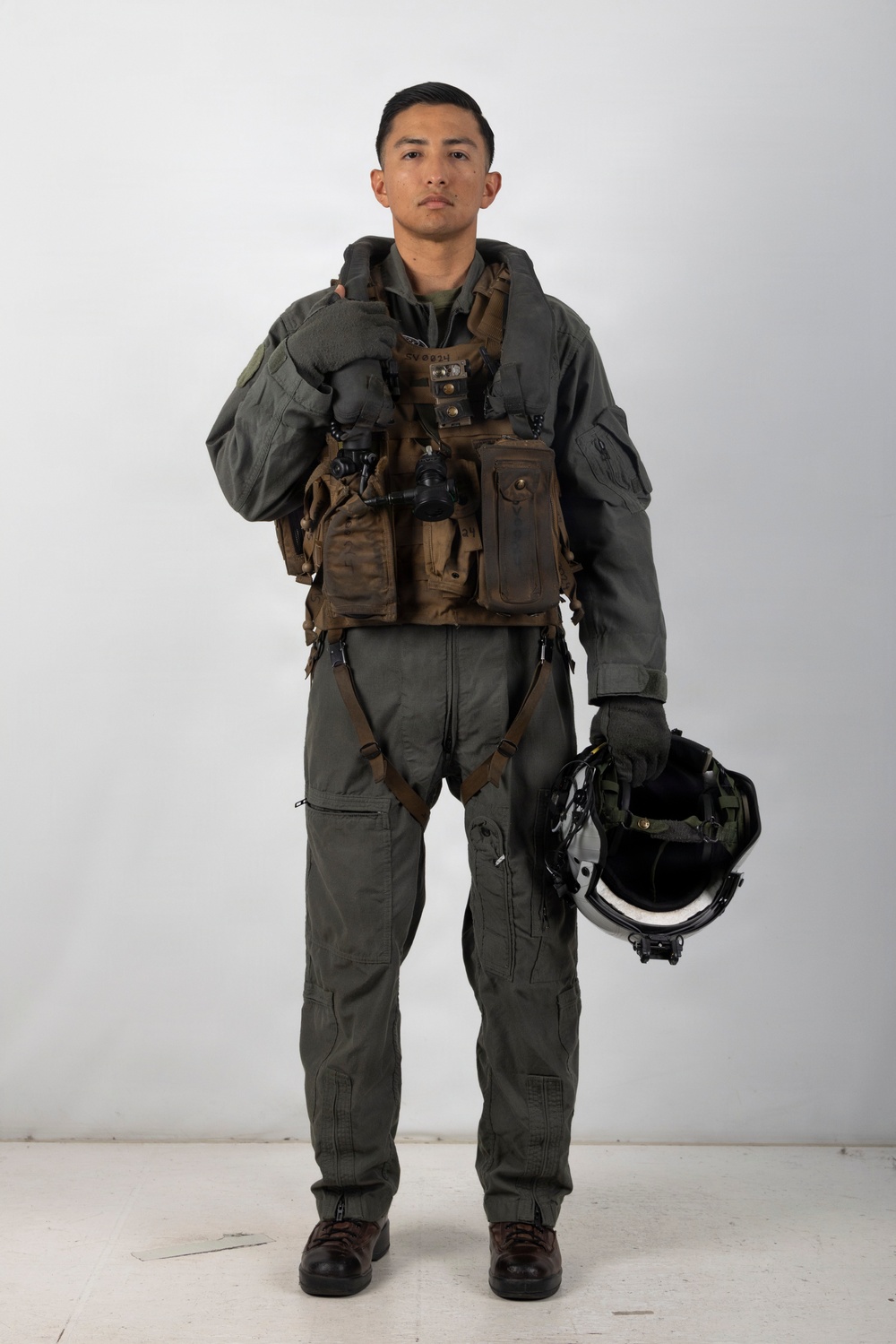 A military aviator stands in a full-body portrait pose against a plain white/light gray studio background. The individual is wearing a complete flight suit ensemble including a survival vest, harness, and is holding a flight helmet at their side. The image appears to be an official or documentary-style photograph.