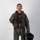 MCAS Marines pose for content 2026 A military aviator stands in a full-body portrait pose against a plain white/light gray studio background. The individual is wearing a complete flight suit ensemble including a survival vest, harness, and is holding a flight helmet at their side. The image appears to be an official or documentary-style photograph.