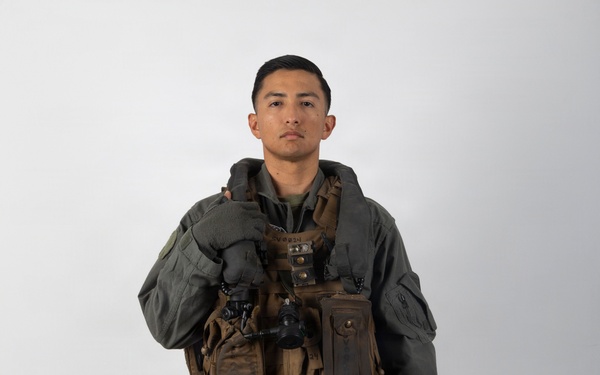 A military aviator stands in a full-body portrait pose against a plain white/light gray studio background. The individual is wearing a complete flight suit ensemble including a survival vest, harness, and is holding a flight helmet at their side. The image appears to be an official or documentary-style photograph.