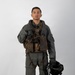 A military aviator stands in a full-body portrait pose against a plain white/light gray studio background. The individual is wearing a complete flight suit ensemble including a survival vest, harness, and is holding a flight helmet at their side. The image appears to be an official or documentary-style photograph.