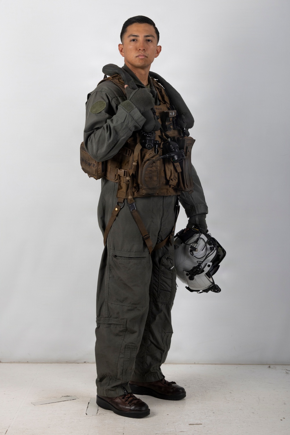 A military aviator is posed in full flight gear against a plain light gray studio background. The subject is wearing an olive drab flight suit with a tactical survival vest, harness, and is holding a flight helmet in one hand. The image appears to be a formal or official portrait-style photograph.