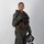 MCAS Marines pose for content 2026 A military aviator is posed in full flight gear against a plain light gray studio background. The subject is wearing an olive drab flight suit with a tactical survival vest, harness, and is holding a flight helmet in one hand. The image appears to be a formal or official portrait-style photograph.
