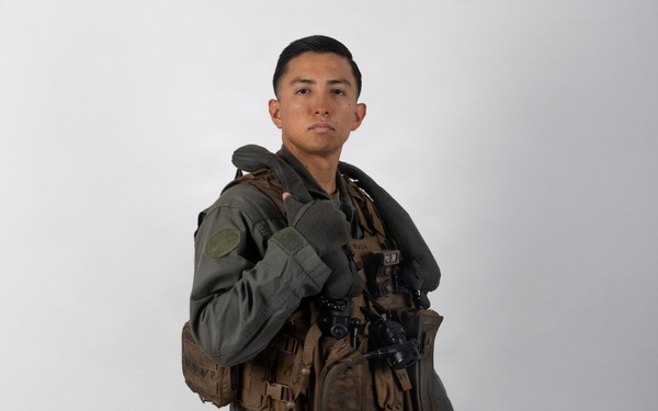 A military aviator is posed in full flight gear against a plain light gray studio background. The subject is wearing an olive drab flight suit with a tactical survival vest, harness, and is holding a flight helmet in one hand. The image appears to be a formal or official portrait-style photograph.