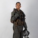A military aviator is posed in full flight gear against a plain light gray studio background. The subject is wearing an olive drab flight suit with a tactical survival vest, harness, and is holding a flight helmet in one hand. The image appears to be a formal or official portrait-style photograph.