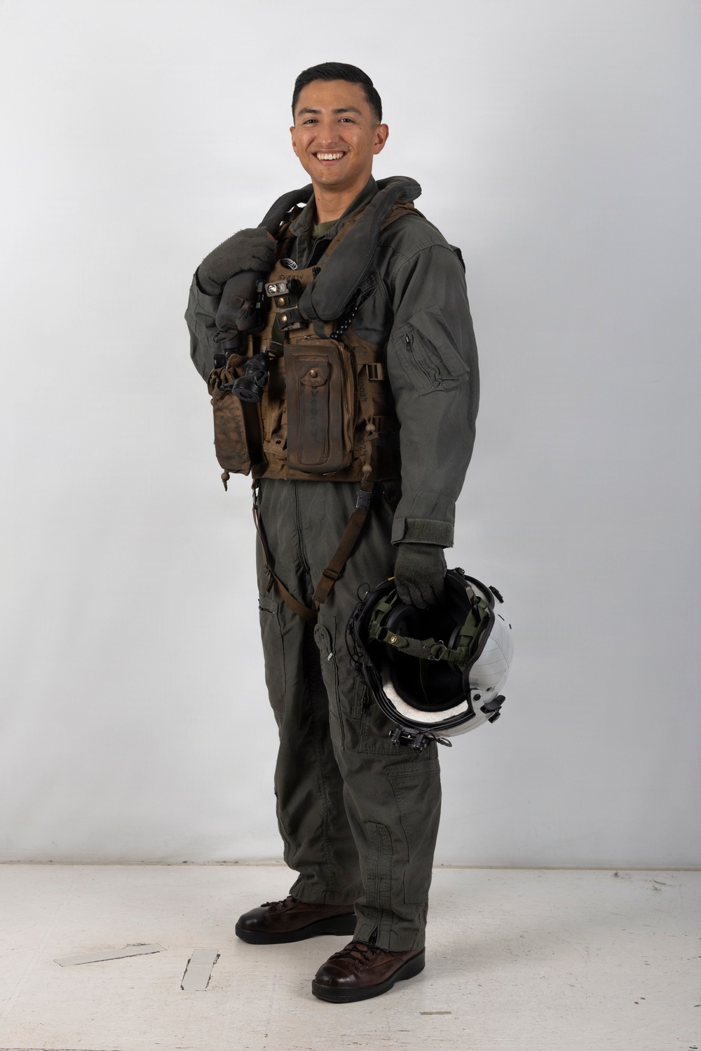 A smiling military aviator poses in full flight gear against a plain white/light gray studio backdrop. The subject is wearing an olive drab flight suit with a survival vest and inflatable collar, and is holding a flight helmet at his side. The image appears to be a formal or promotional portrait photograph.