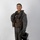 MCAS Marines pose for content 2026 A smiling military aviator poses in full flight gear against a plain white/light gray studio backdrop. The subject is wearing an olive drab flight suit with a survival vest and inflatable collar, and is holding a flight helmet at his side. The image appears to be a formal or promotional portrait photograph.