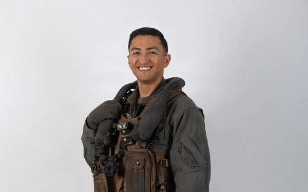 A smiling military aviator poses in full flight gear against a plain white/light gray studio backdrop. The subject is wearing an olive drab flight suit with a survival vest and inflatable collar, and is holding a flight helmet at his side. The image appears to be a formal or promotional portrait photograph.