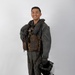 A smiling military aviator poses in full flight gear against a plain white/light gray studio backdrop. The subject is wearing an olive drab flight suit with a survival vest and inflatable collar, and is holding a flight helmet at his side. The image appears to be a formal or promotional portrait photograph.