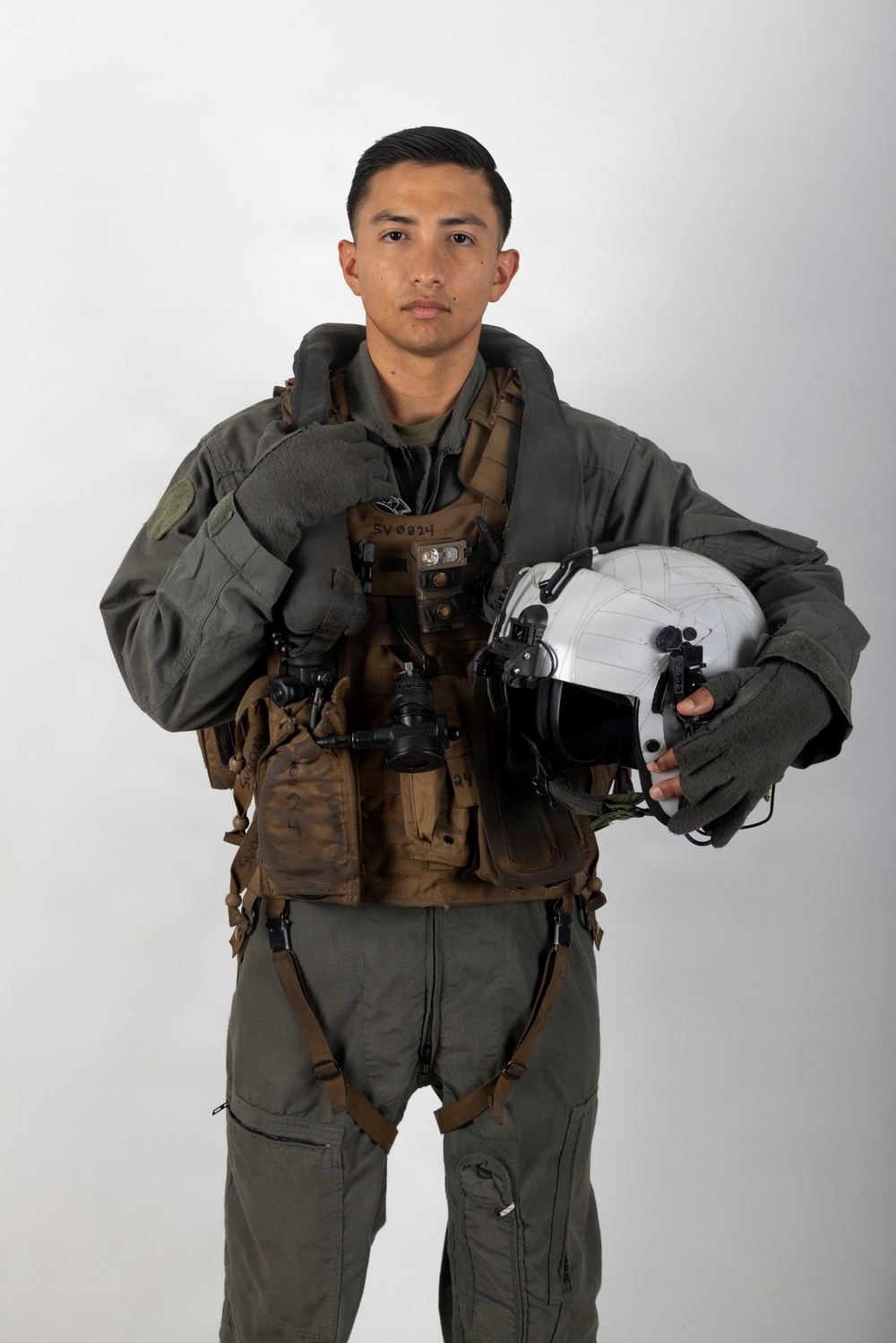 A military aviator poses for a formal portrait against a neutral gray-white background. The individual is wearing a full flight suit ensemble including a green flight suit, survival vest, harness system, and gloves, while holding a white flight helmet. The image appears to be an official or professional military portrait photograph.