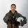 MCAS Marines pose for content 2026 A military aviator poses for a formal portrait against a neutral gray-white background. The individual is wearing a full flight suit ensemble including a green flight suit, survival vest, harness system, and gloves, while holding a white flight helmet. The image appears to be an official or professional military portrait photograph.