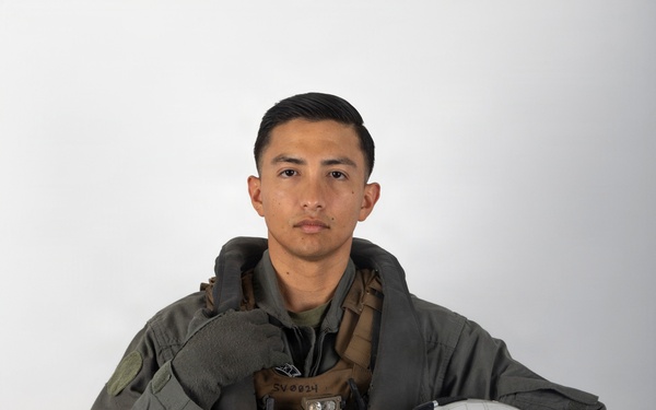 A military aviator poses for a formal portrait against a neutral gray-white background. The individual is wearing a full flight suit ensemble including a green flight suit, survival vest, harness system, and gloves, while holding a white flight helmet. The image appears to be an official or professional military portrait photograph.