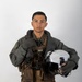 A military aviator poses for a formal portrait against a neutral gray-white background. The individual is wearing a full flight suit ensemble including a green flight suit, survival vest, harness system, and gloves, while holding a white flight helmet. The image appears to be an official or professional military portrait photograph.