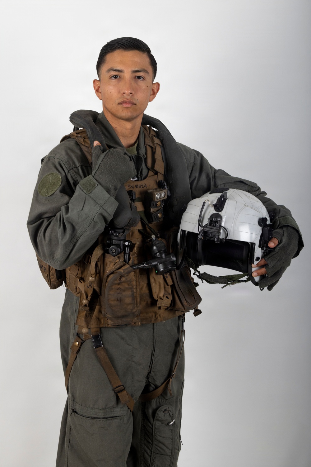A young male military aviator poses against a neutral gray studio background in full flight gear. He is holding a white flight helmet in his right hand and wearing a green flight suit with a tactical survival vest. The image appears to be an official or professional portrait photograph.