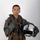 MCAS Marines pose for content 2026 A young male military aviator poses against a neutral gray studio background in full flight gear. He is holding a white flight helmet in his right hand and wearing a green flight suit with a tactical survival vest. The image appears to be an official or professional portrait photograph.