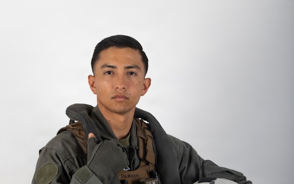 A young male military aviator poses against a neutral gray studio background in full flight gear. He is holding a white flight helmet in his right hand and wearing a green flight suit with a tactical survival vest. The image appears to be an official or professional portrait photograph.