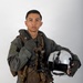 A young male military aviator poses against a neutral gray studio background in full flight gear. He is holding a white flight helmet in his right hand and wearing a green flight suit with a tactical survival vest. The image appears to be an official or professional portrait photograph.