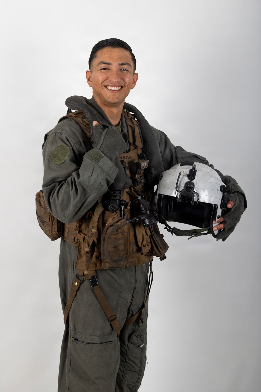 A smiling young military aviator poses in a professional portrait-style photograph against a neutral white/grey background. He is wearing a full flight suit with tactical gear and harness, holding a white flight helmet in one hand while giving a thumbs-up with the other. The image appears to be an official or semi-official military portrait.