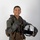 MCAS Marines pose for content 2026 A smiling young military aviator poses in a professional portrait-style photograph against a neutral white/grey background. He is wearing a full flight suit with tactical gear and harness, holding a white flight helmet in one hand while giving a thumbs-up with the other. The image appears to be an official or semi-official military portrait.