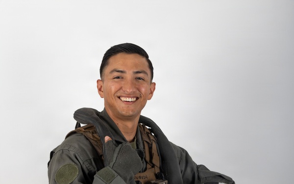 A smiling young military aviator poses in a professional portrait-style photograph against a neutral white/grey background. He is wearing a full flight suit with tactical gear and harness, holding a white flight helmet in one hand while giving a thumbs-up with the other. The image appears to be an official or semi-official military portrait.