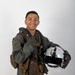 A smiling young military aviator poses in a professional portrait-style photograph against a neutral white/grey background. He is wearing a full flight suit with tactical gear and harness, holding a white flight helmet in one hand while giving a thumbs-up with the other. The image appears to be an official or semi-official military portrait.