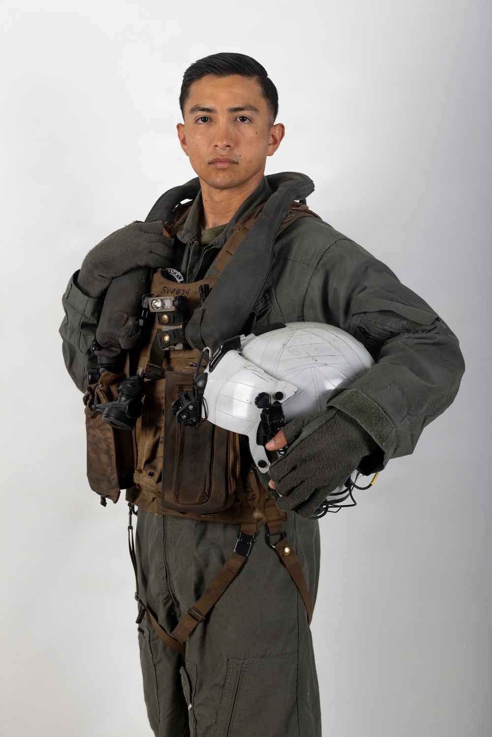 A military aviator poses against a neutral white/gray background in a formal portrait-style photograph. The individual is wearing a full flight suit ensemble including a survival vest, harness system, and is holding a white flight helmet. The image appears to be an official military portrait.