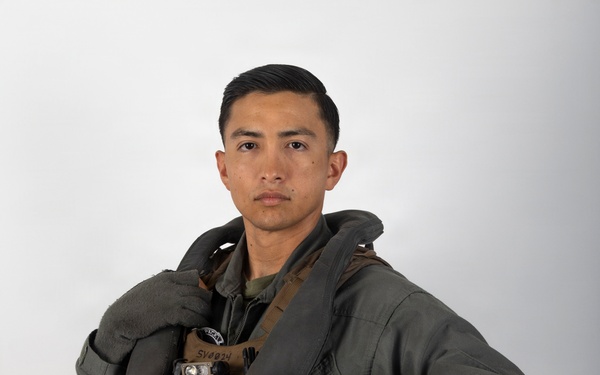 A military aviator poses against a neutral white/gray background in a formal portrait-style photograph. The individual is wearing a full flight suit ensemble including a survival vest, harness system, and is holding a white flight helmet. The image appears to be an official military portrait.