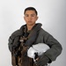 A military aviator poses against a neutral white/gray background in a formal portrait-style photograph. The individual is wearing a full flight suit ensemble including a survival vest, harness system, and is holding a white flight helmet. The image appears to be an official military portrait.