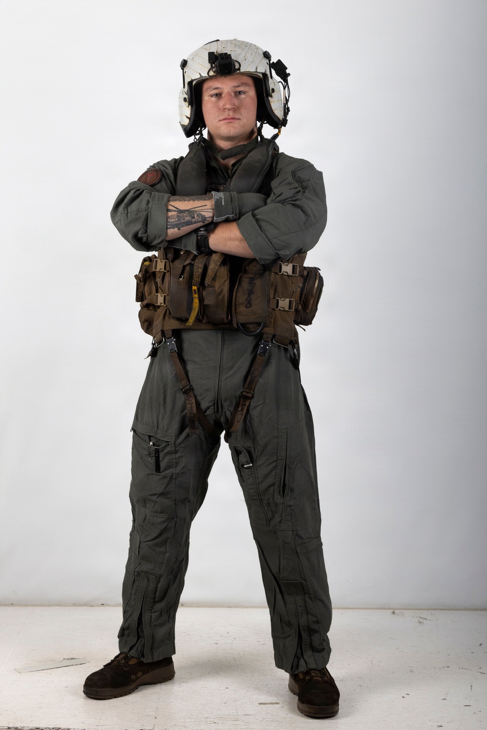 A military aviator stands in a full-body portrait pose against a white studio background with arms crossed. The individual is wearing a complete flight suit ensemble including a flight helmet, survival vest, and harness system. The image appears to be a professional or official military portrait photograph.