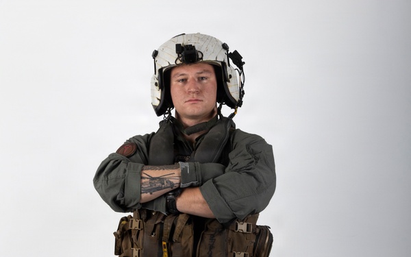 A military aviator stands in a full-body portrait pose against a white studio background with arms crossed. The individual is wearing a complete flight suit ensemble including a flight helmet, survival vest, and harness system. The image appears to be a professional or official military portrait photograph.