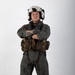 A military aviator stands in a full-body portrait pose against a white studio background with arms crossed. The individual is wearing a complete flight suit ensemble including a flight helmet, survival vest, and harness system. The image appears to be a professional or official military portrait photograph.