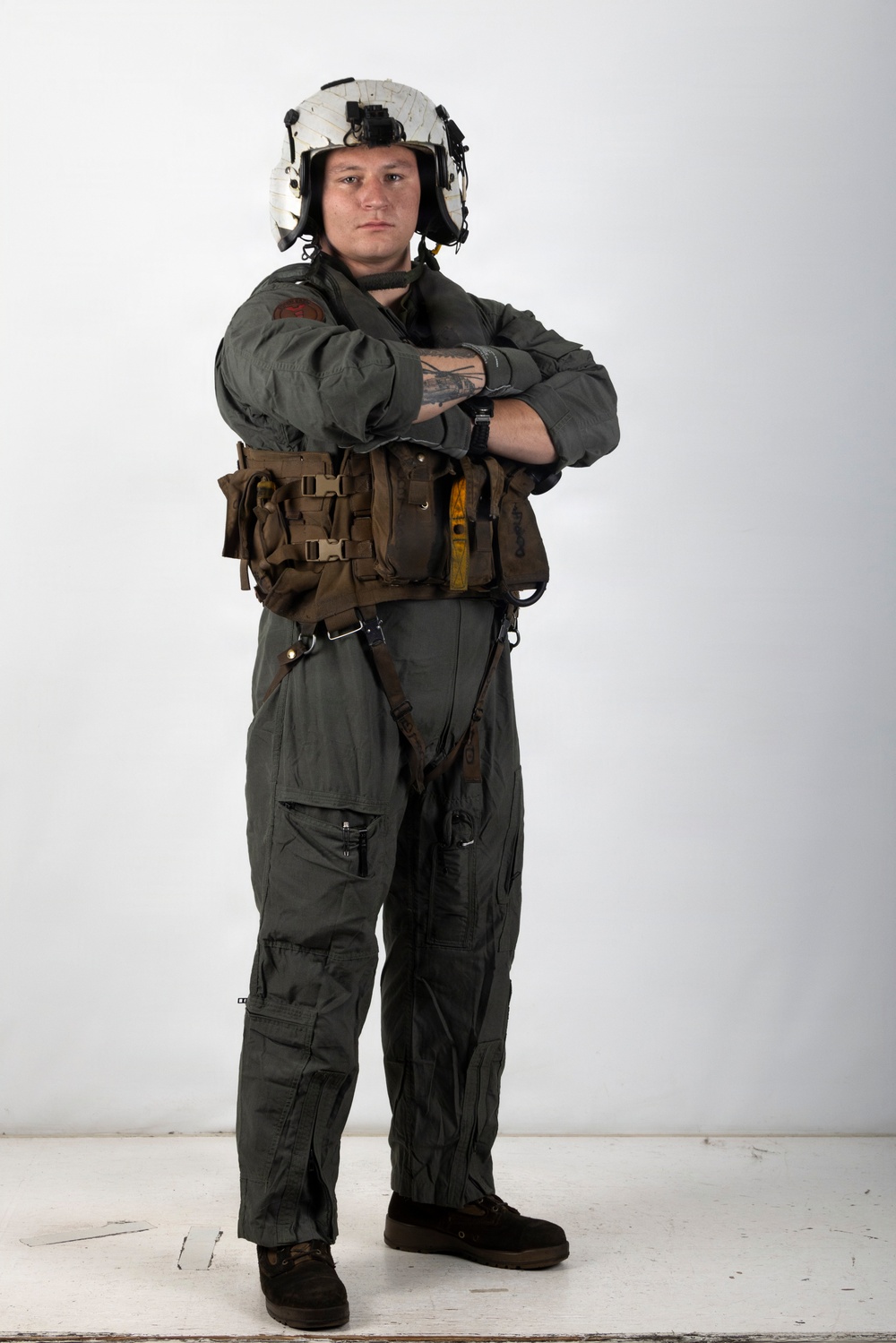 A military aviator stands in a full-body portrait pose against a white studio background with arms crossed. The individual is wearing a flight suit with survival/tactical gear and a white flight helmet. The image appears to be a professional or official-style portrait photograph.