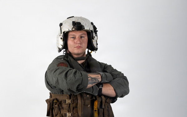 A military aviator stands in a full-body portrait pose against a white studio background with arms crossed. The individual is wearing a flight suit with survival/tactical gear and a white flight helmet. The image appears to be a professional or official-style portrait photograph.