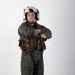 A military aviator stands in a full-body portrait pose against a white studio background with arms crossed. The individual is wearing a flight suit with survival/tactical gear and a white flight helmet. The image appears to be a professional or official-style portrait photograph.