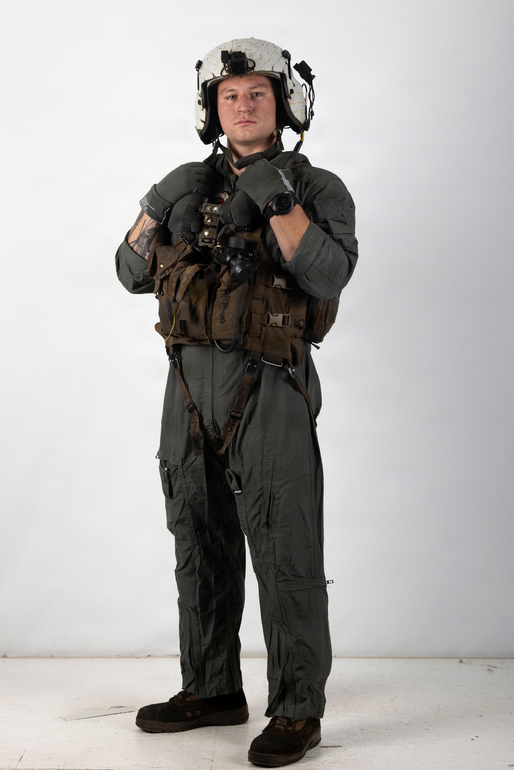 A military aviator stands in a full flight suit ensemble against a plain white studio background. The individual is wearing complete flight gear including a helmet, survival vest, harness, and gloves, posed in a formal standing position. The image appears to be a professional or official documentation photograph.