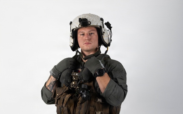 A military aviator stands in a full flight suit ensemble against a plain white studio background. The individual is wearing complete flight gear including a helmet, survival vest, harness, and gloves, posed in a formal standing position. The image appears to be a professional or official documentation photograph.