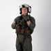 A military aviator stands in a full flight suit ensemble against a plain white studio background. The individual is wearing complete flight gear including a helmet, survival vest, harness, and gloves, posed in a formal standing position. The image appears to be a professional or official documentation photograph.