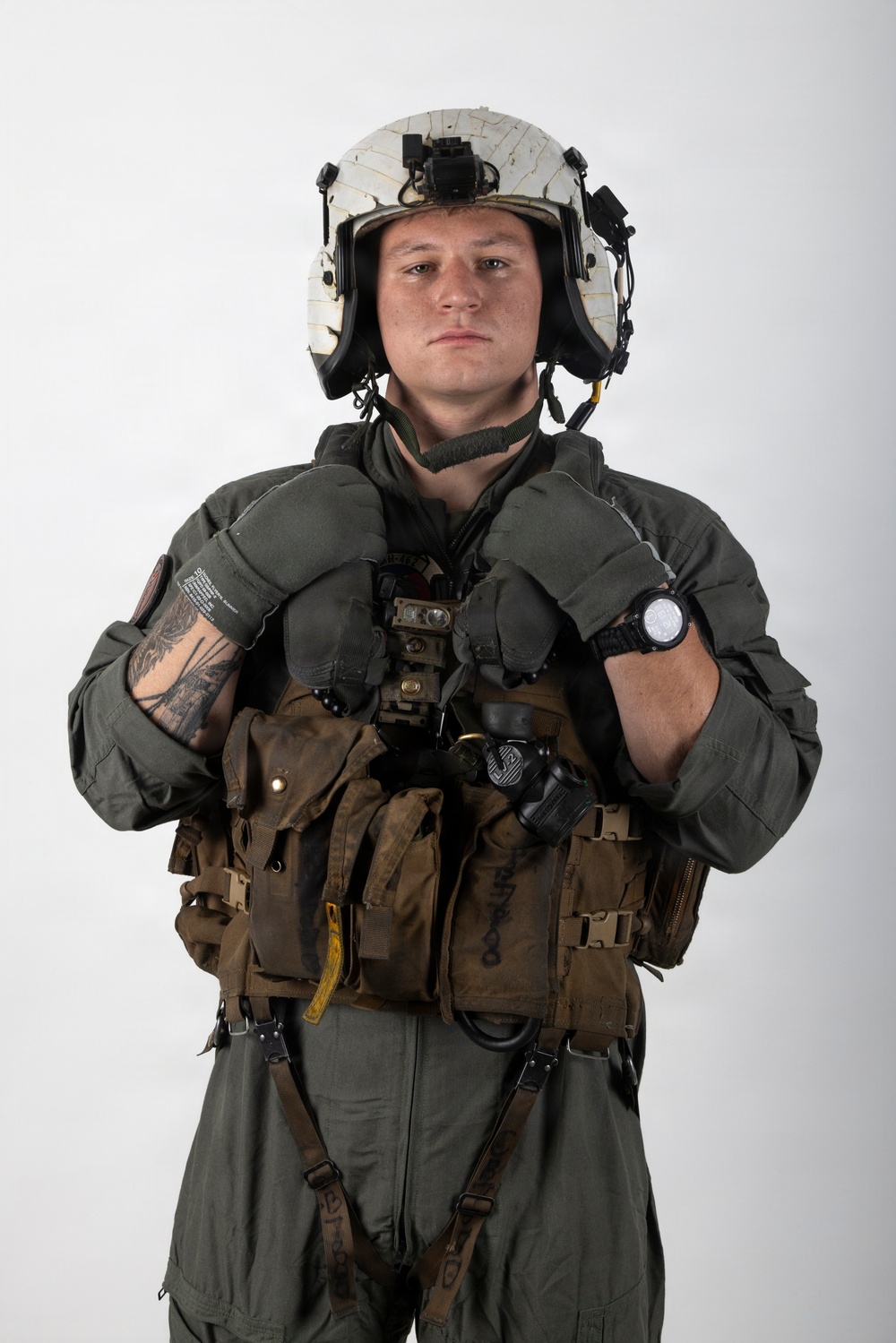 A military aviator or aircrew member is photographed against a plain white background in a formal portrait-style image. The individual is wearing full flight gear including a flight suit, survival vest, flight helmet, and gloves. The image appears to be an official or semi-official military portrait.