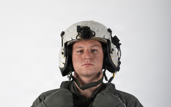 A military aviator or aircrew member is photographed against a plain white background in a formal portrait-style image. The individual is wearing full flight gear including a flight suit, survival vest, flight helmet, and gloves. The image appears to be an official or semi-official military portrait.