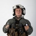 A military aviator or aircrew member is photographed against a plain white background in a formal portrait-style image. The individual is wearing full flight gear including a flight suit, survival vest, flight helmet, and gloves. The image appears to be an official or semi-official military portrait.