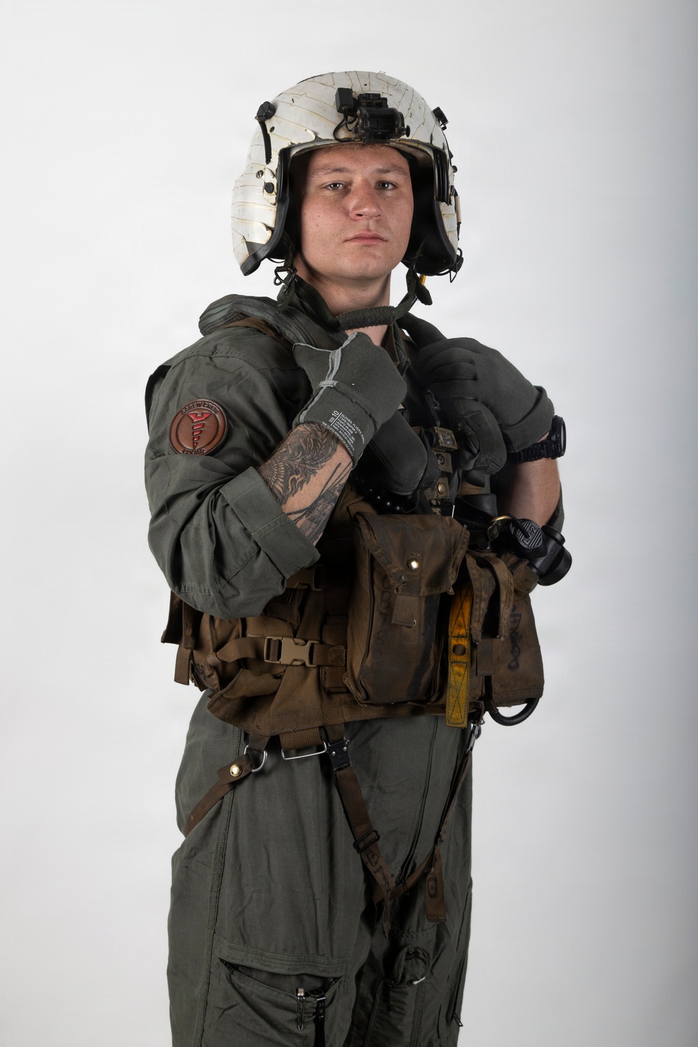 A young male in full military flight crew gear poses against a plain white/grey studio background. He is wearing a flight suit, survival vest, flight helmet with night vision mount, and tactical gloves. The image appears to be a professional portrait or documentation photograph.